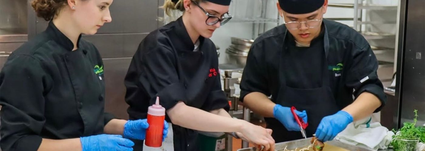 Three young chefs in black chef coats and hats work together in a professional kitchen. The recent UFV culinary grads are focused and collaborative as they prepare a meal, surrounded by stainless steel equipment and ingredients.