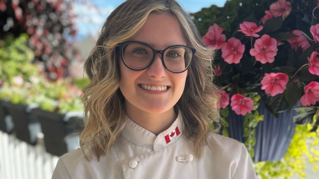 Outdoor headshot of a young chef and recent UFV culinary graduate, smiling confidently at the camera. She wears a chef’s coat and stands in natural light, with pink flowers in the background.