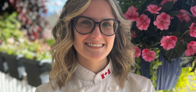 Outdoor headshot of a young chef and recent UFV culinary graduate, smiling confidently at the camera. She wears a chef’s coat and stands in natural light, with pink flowers in the background.